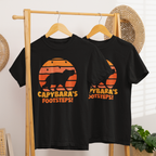 Áo thun đen in hình capybara silhouette trên nền hoàng hôn cam kèm dấu chân, chữ “Capybara’s Footsteps!” nổi bật