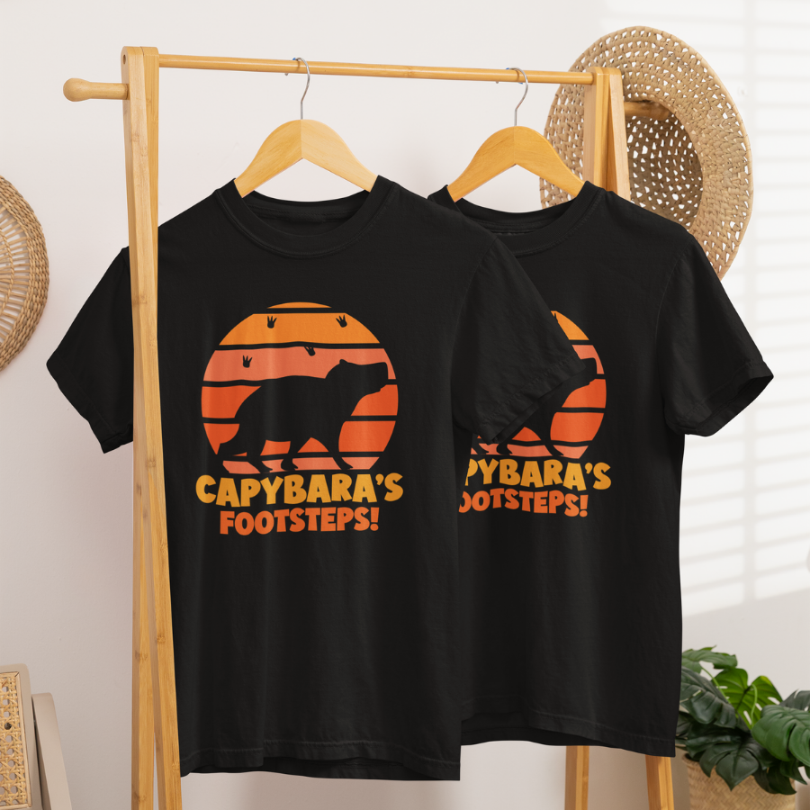 Áo thun đen in hình capybara silhouette trên nền hoàng hôn cam kèm dấu chân, chữ “Capybara’s Footsteps!” nổi bật