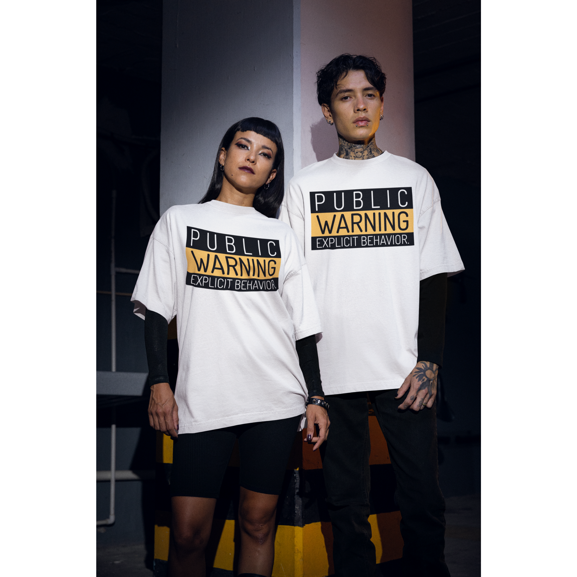 Hai người mặc áo thun trắng in chữ “PUBLIC WARNING: EXPLICIT BEHAVIOR” phong cách streetwear cá tính.